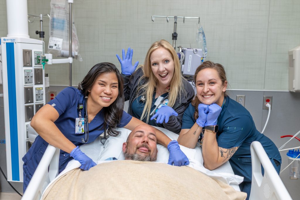 Three female nurses smiling and standing behind a patient in a bed.