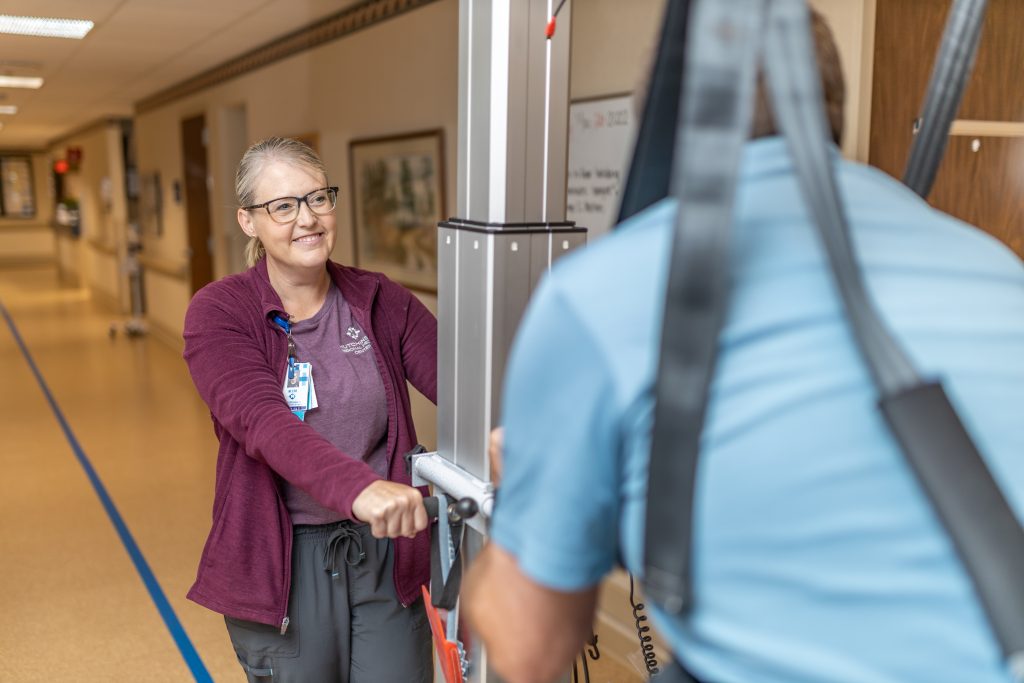 Female nurse in a dark purple jacket, helping a patient walk down the hallway